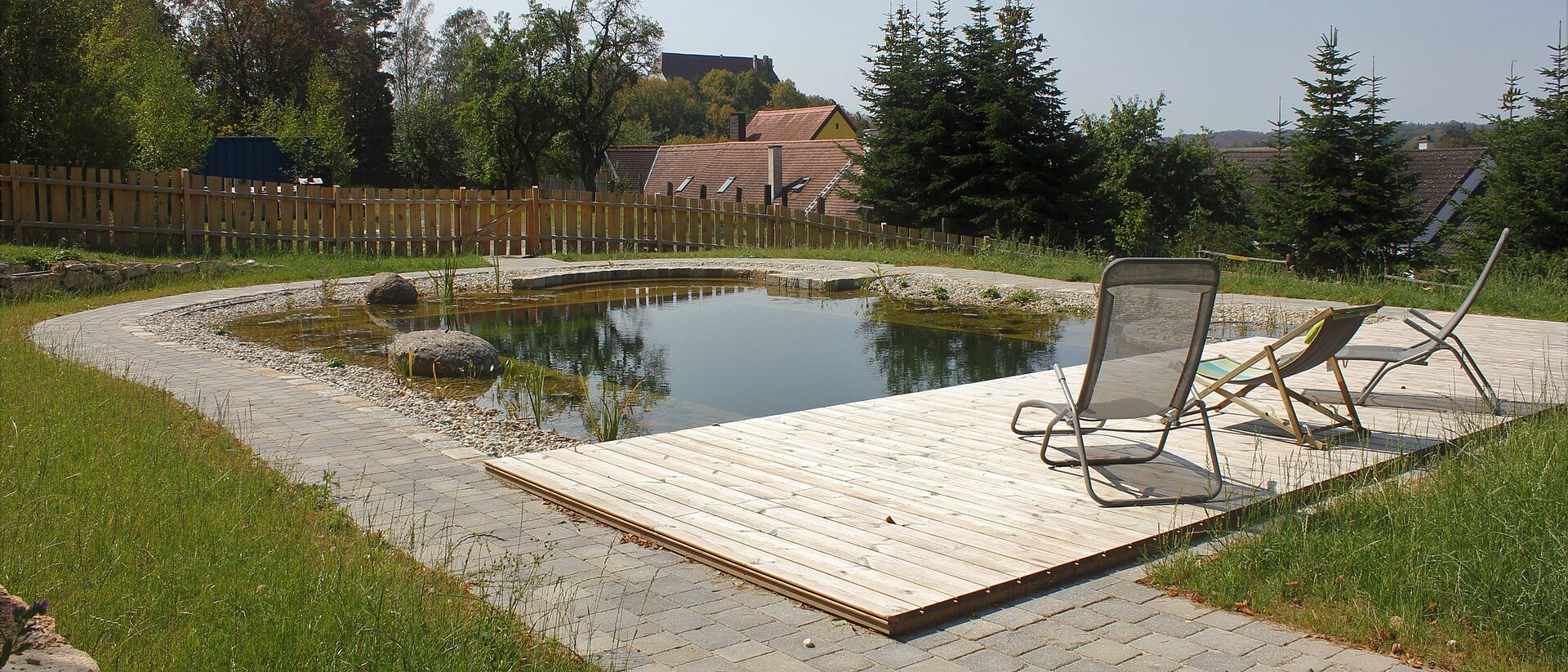 Naturpool mit Holzsteg und Sonnenliegen, im Hintergrund das Forsthaus
